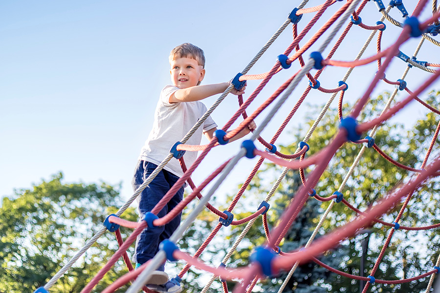 Little boy wearing a white t-shirt and blue jeans climbing up a red and blue climbing net