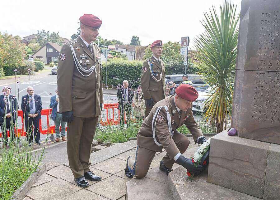 Military personnel in uniform, with one kneeling to lay a wreath at a Polish memorial stone, surrounded by a small group of people in an outdoor setting