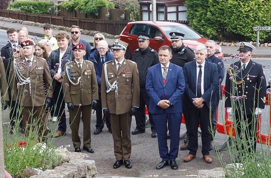 Group of people, including military personnel in uniform, police officers, and civilians, gathered outdoors near Polish memorial 